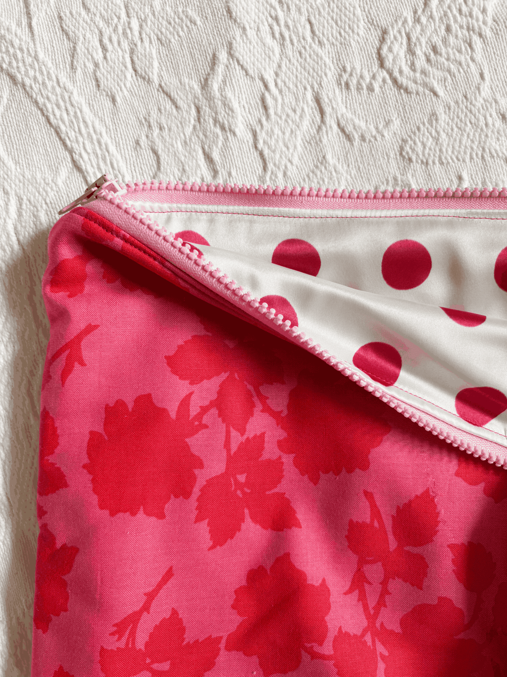 A close-up of a zippered laptop sleeve, showing a bright pink floral exterior and a white interior with pink polka dots, set against a textured off-white fabric background.