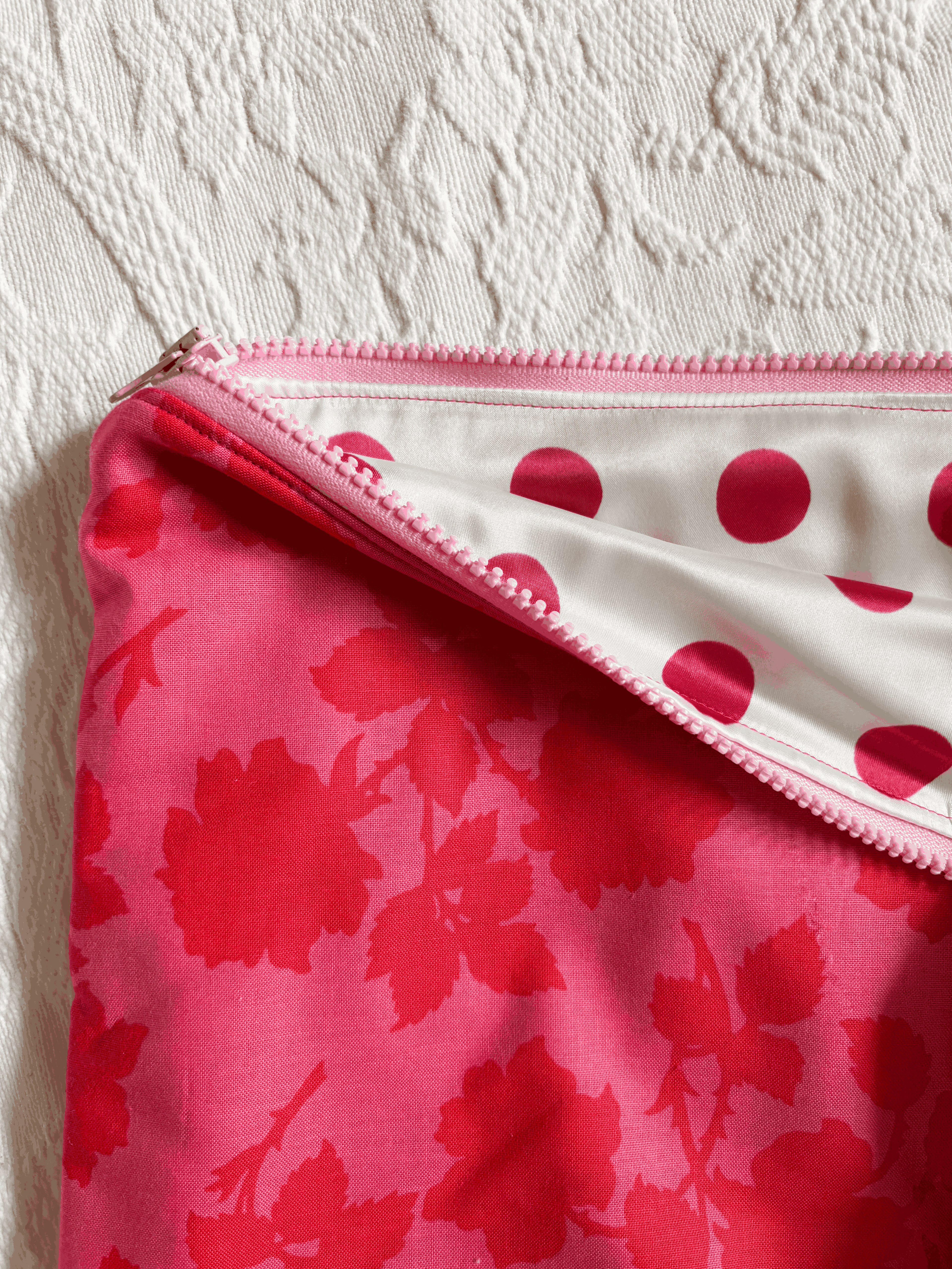 A close-up of a zippered laptop sleeve, showing a bright pink floral exterior and a white interior with pink polka dots, set against a textured off-white fabric background.