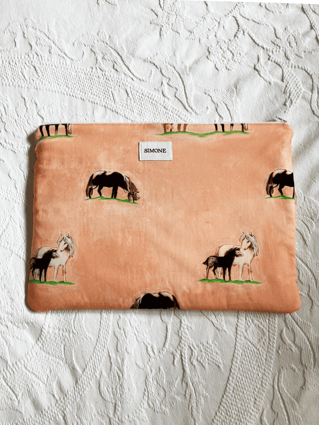 A zippered laptop sleeve in a peach-colored fabric, printed with black and white horses on a green grass background. A small white label on the front reads 'SIMONE'.