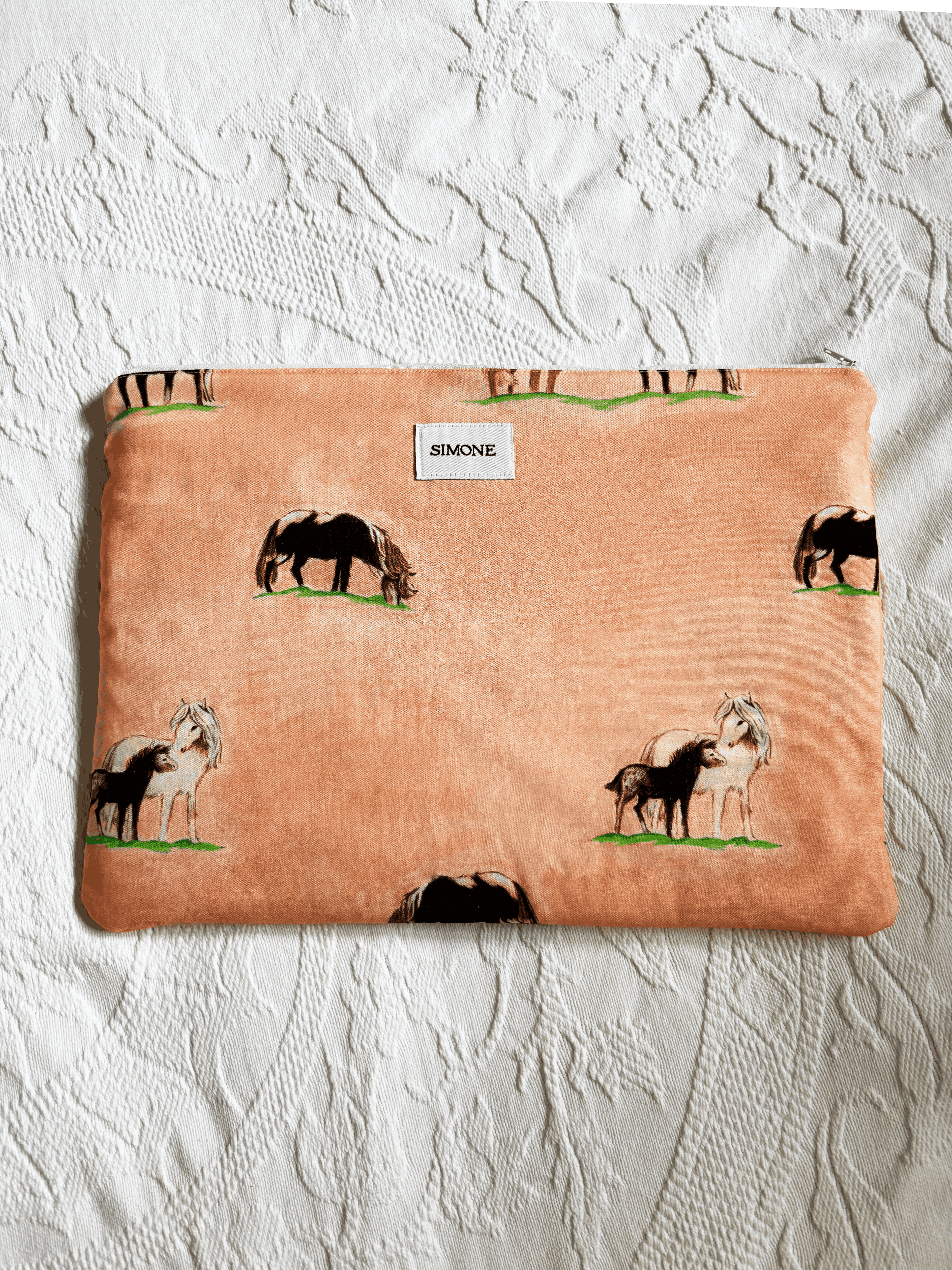 A zippered laptop sleeve in a peach-colored fabric, printed with black and white horses on a green grass background. A small white label on the front reads 'SIMONE'.