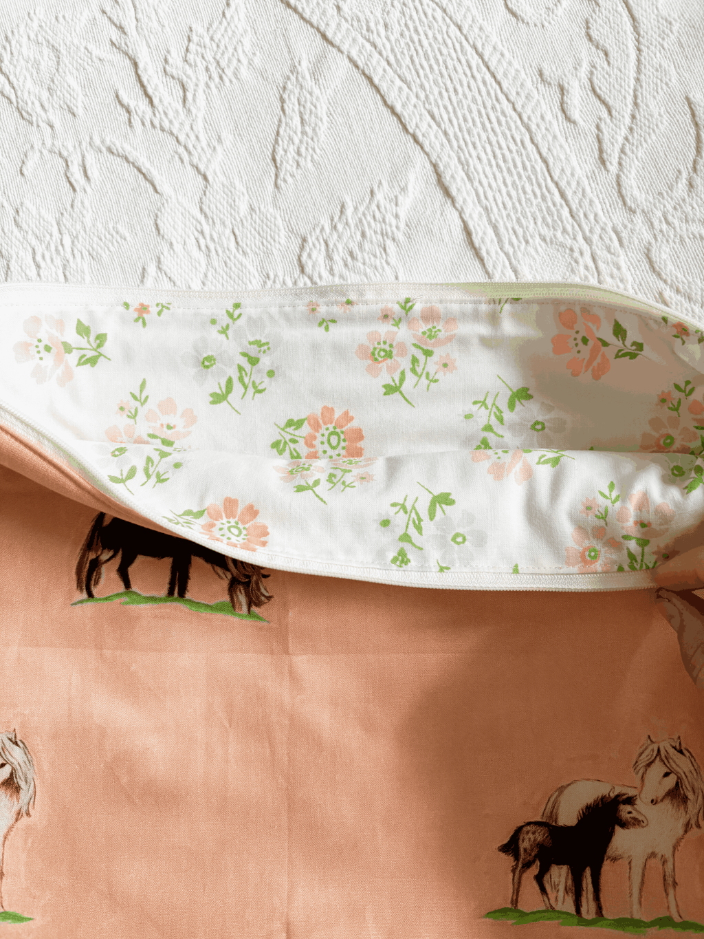 A zippered laptop sleeve in a peach-colored fabric, printed with black and white horses on a green grass background. The interior is white with flowers. 