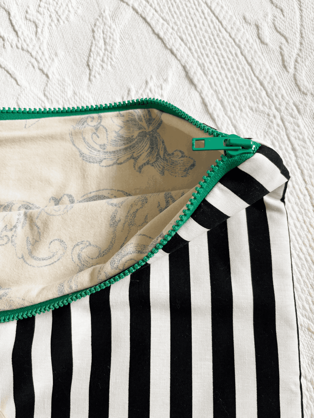 Close-up of zippered laptop sleeve with black and white vertical stripes, a green zipper, and a cream and blue toile lining, laid flat on a white textured surface.