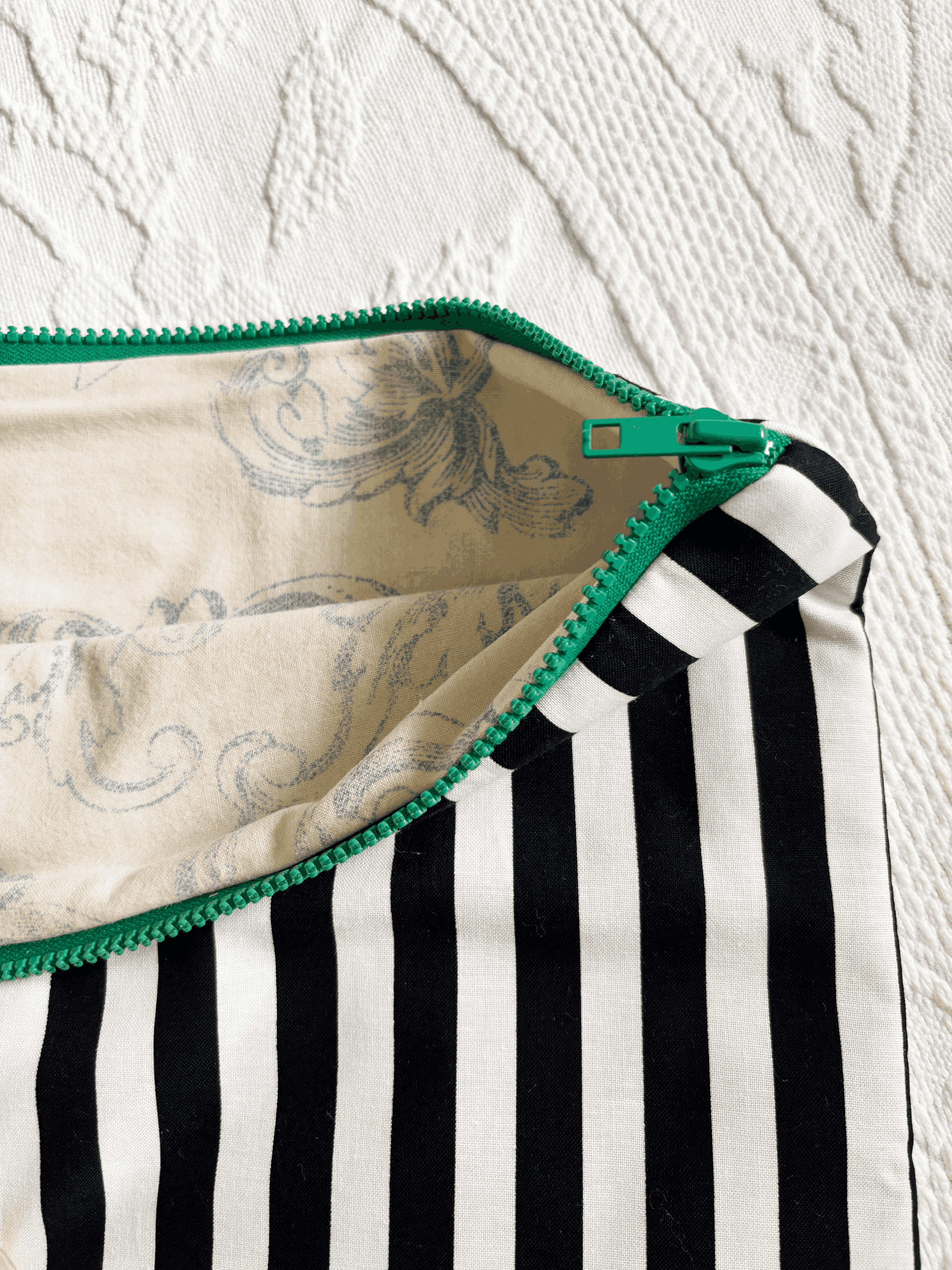 Close-up of zippered laptop sleeve with black and white vertical stripes, a green zipper, and a cream and blue toile lining, laid flat on a white textured surface.