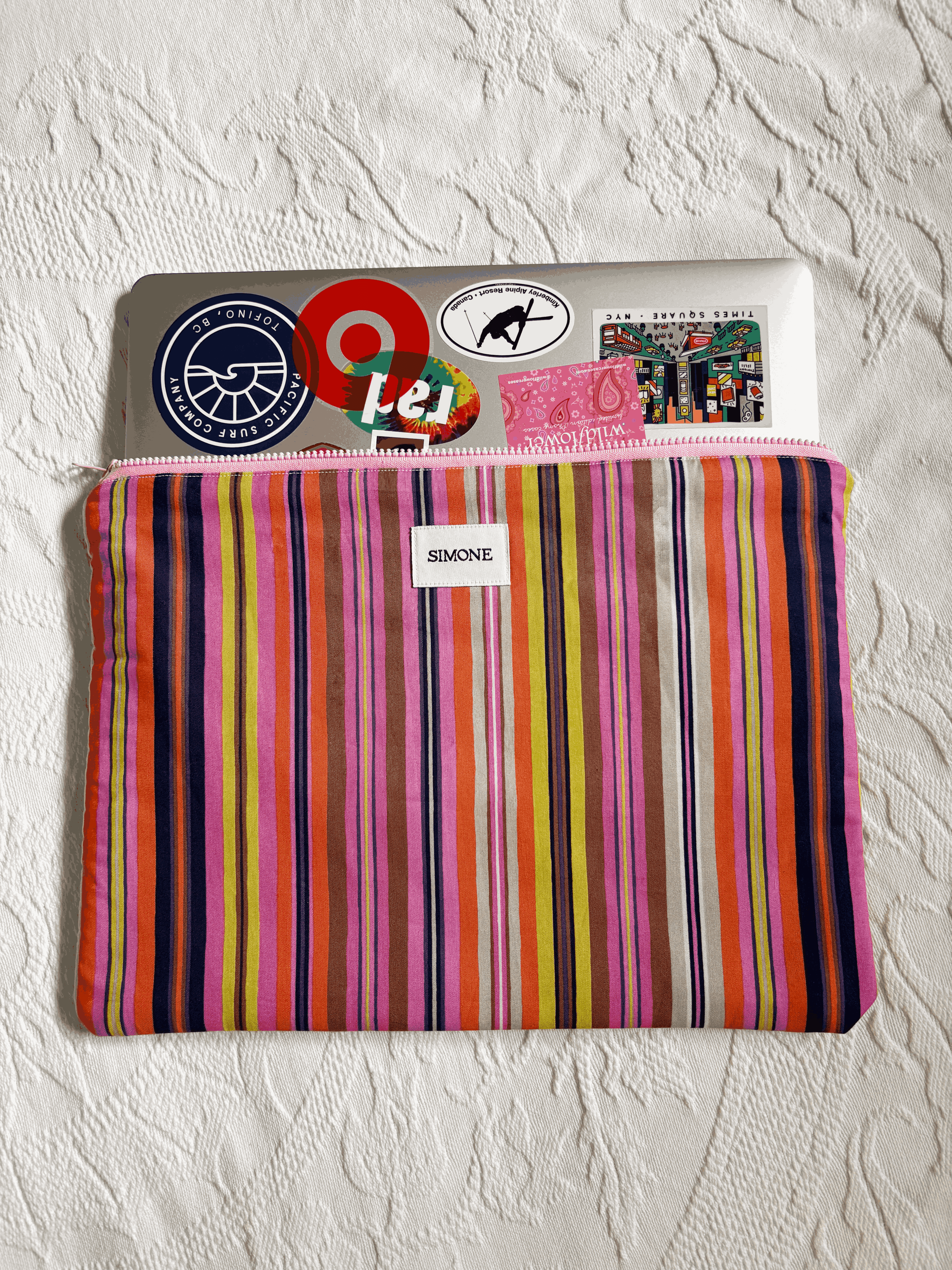 A colorful (pink, yellow, orange, brown, blue) striped fabric pouch with a zipper closure, featuring a white label that reads 'SIMONE' and holding a laptop covered in stickers, placed on a textured white fabric background.