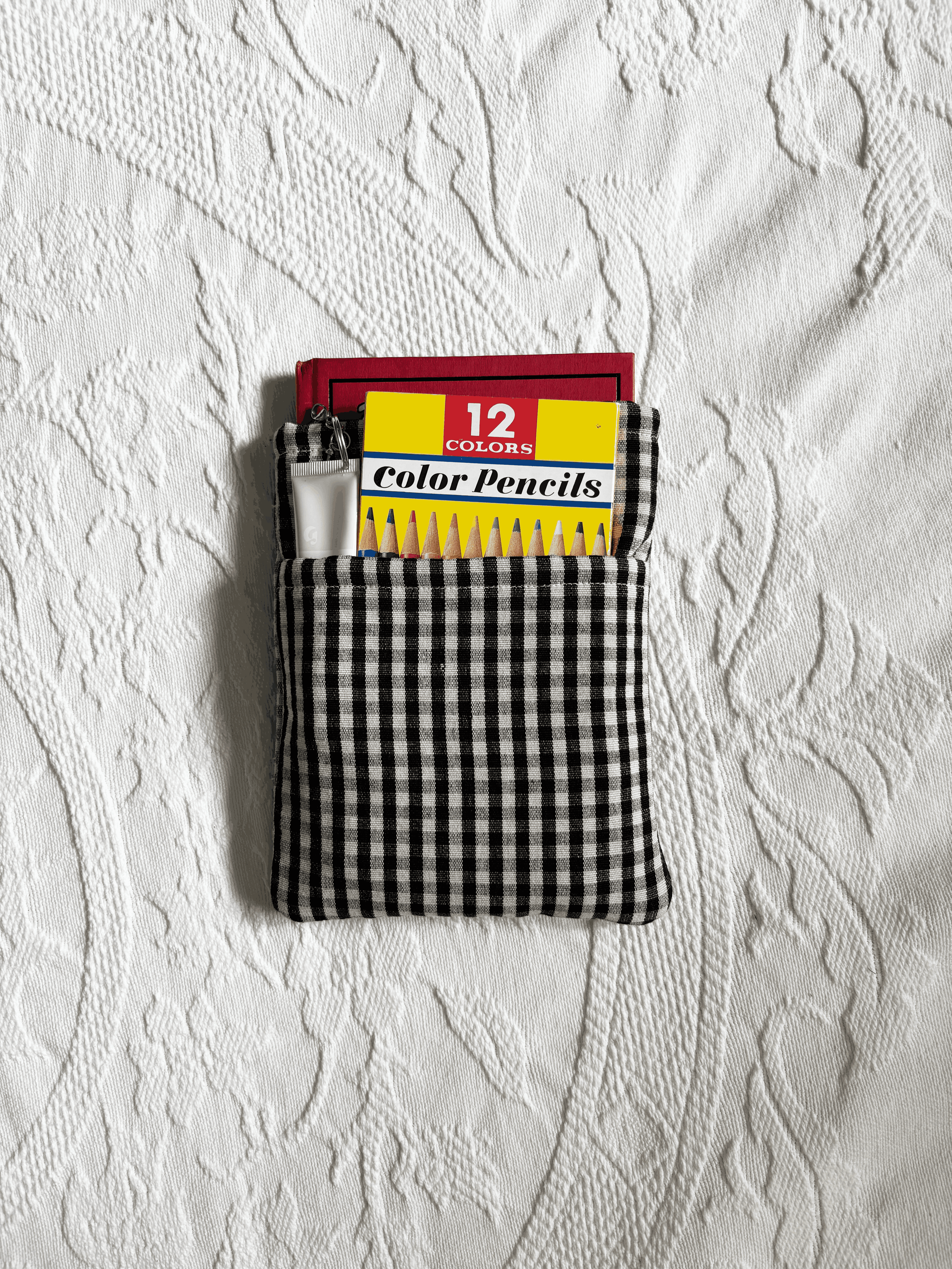 A black and white gingham fabric book sleeve laid flat on a white textured fabric background. The main compartment has a small hardcover book and the top pocket has 12 coloured pencils and a lipbalm.
