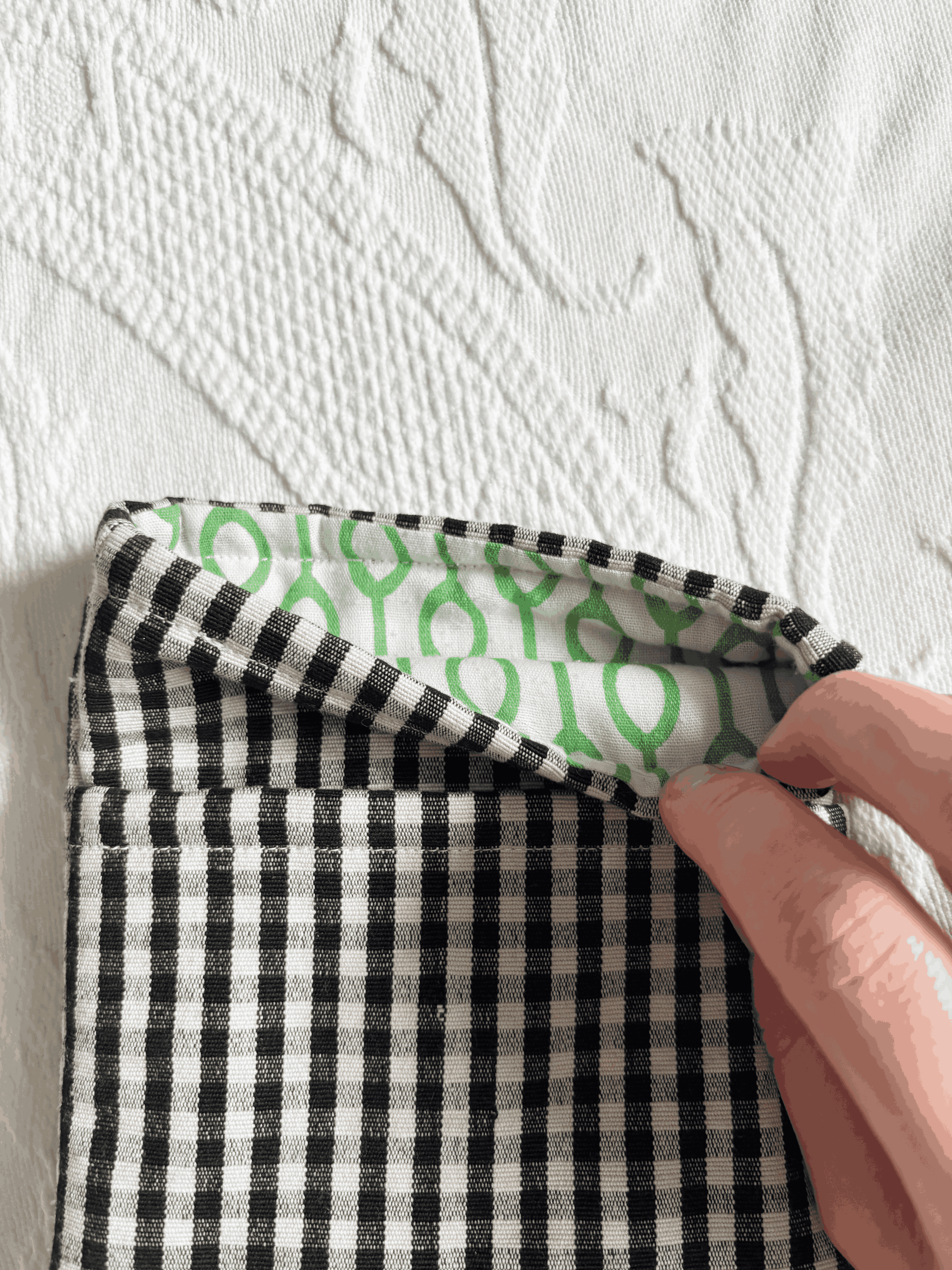 A close-up of a black and white gingham book sleeve being held open by a hand, showing a green patterned lining on a textured white background.