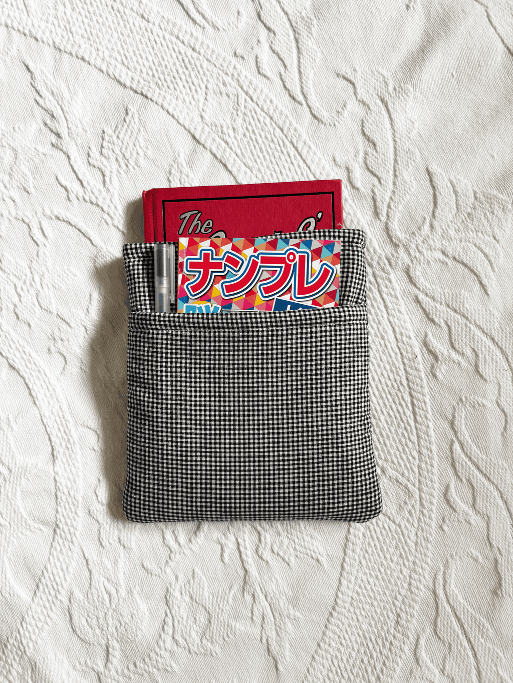 A small black-and-white gingham book sleeeve holding a red notebook, pen, and a colorful magazine, placed on a white textured blanket.