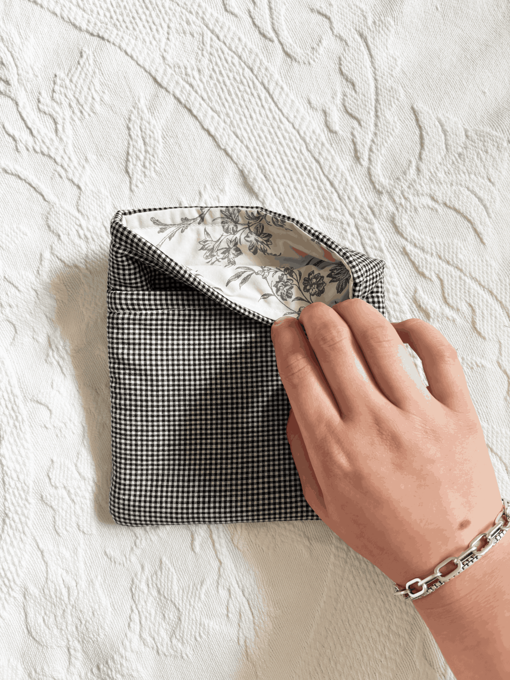 A small black and white gingham book sleeve with a floral-patterned lining, held open by a hand wearing a silver chain bracelet, set against a textured white lace background.