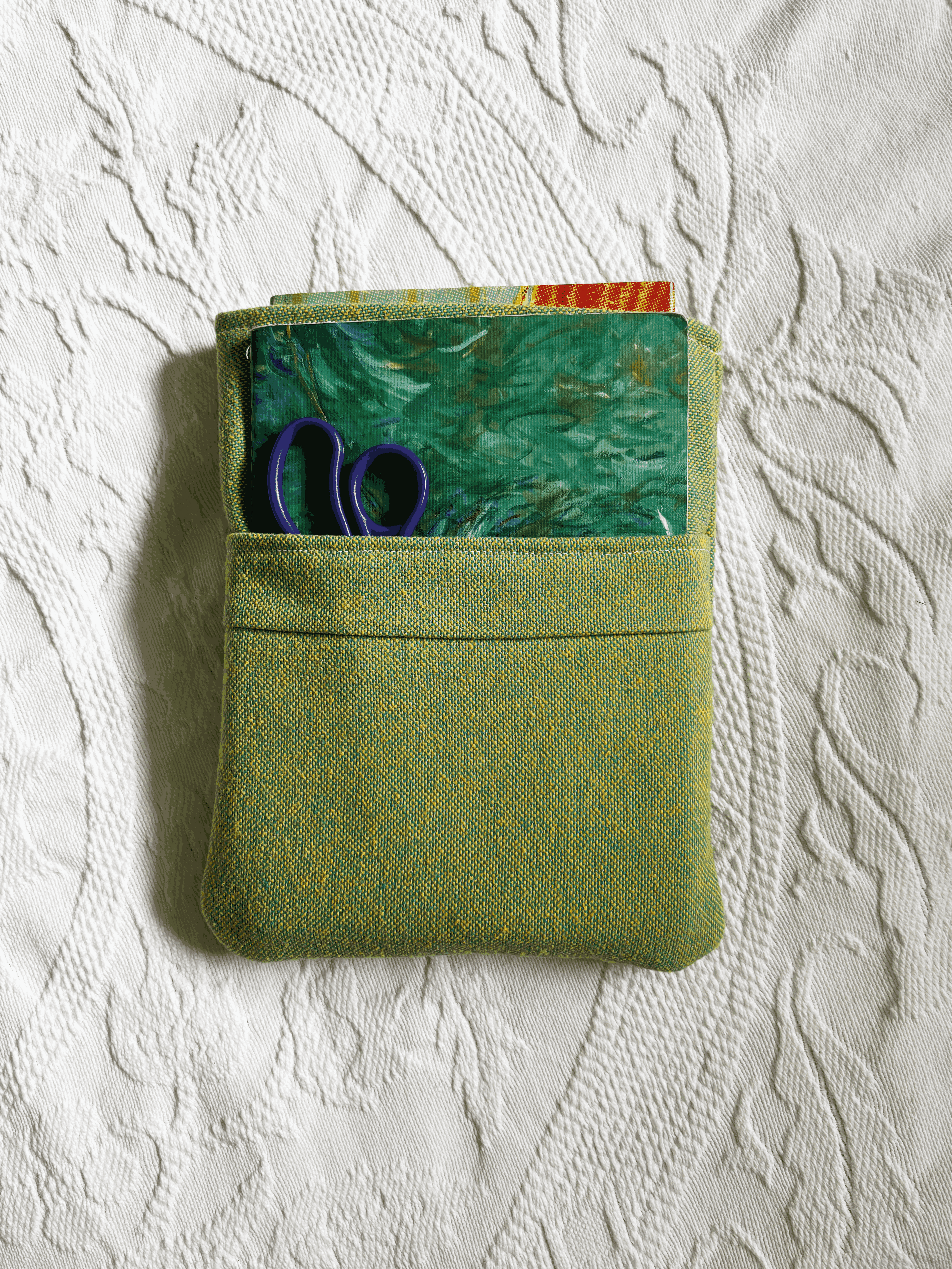 A small green book sleeve with a top pocket, shown on a white textured fabric background. A hardcover book is peeking out of the main compartment while a notebook and pair of scissors is tucked into the front pocket.