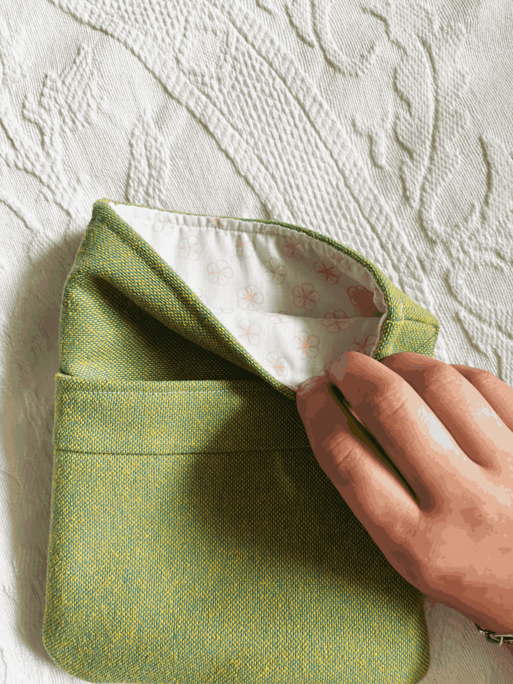 A small green book sleeve with a top pocket, shown on a white textured fabric background. The lining fabric is white with small pink flowers.