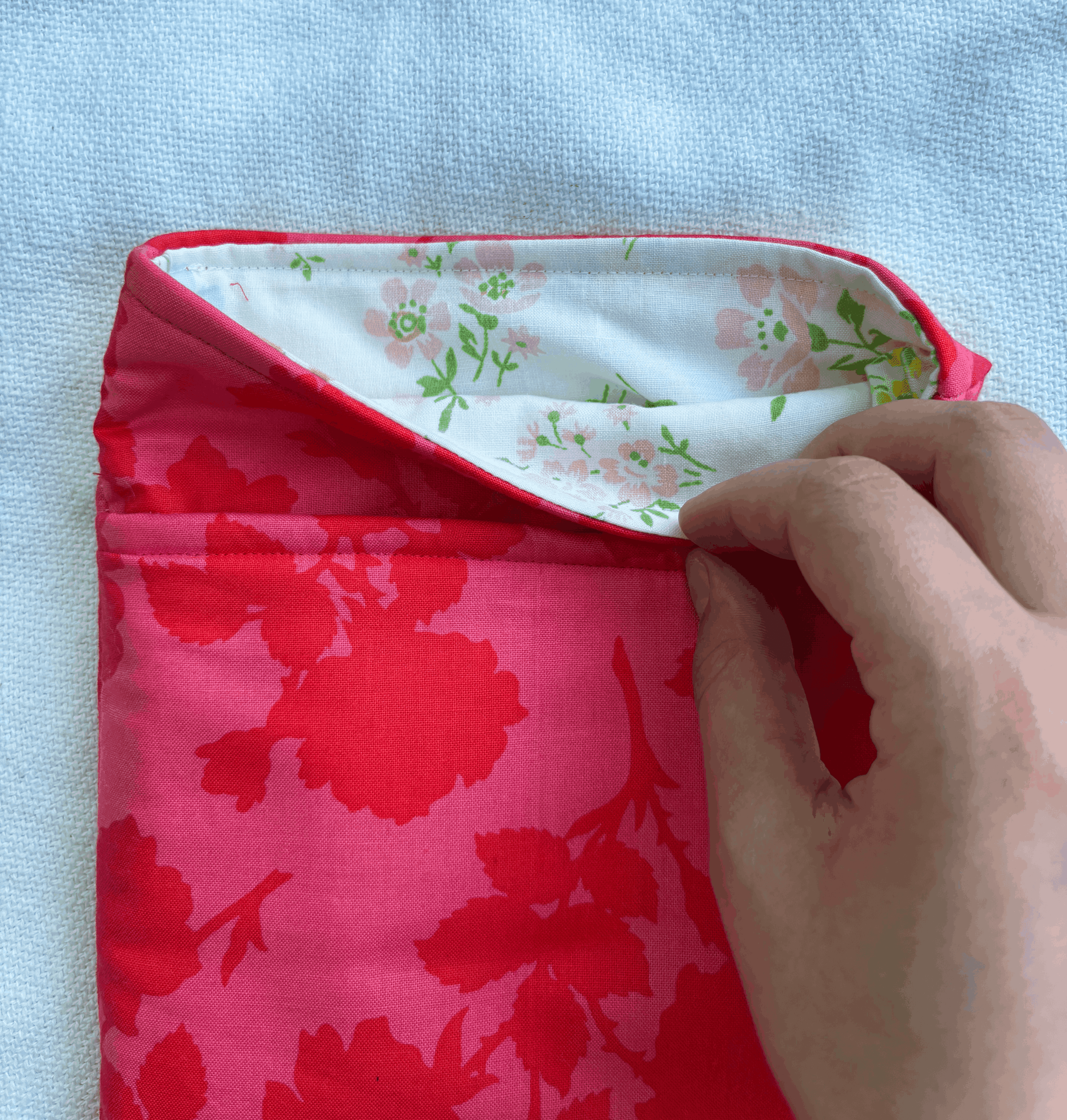 A close-up of a book sleeve with a bright pink floral exterior and a white floral lining, being held open by a person's hand against a light blue textured background.